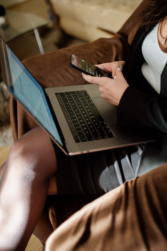 Young businesswoman with curly hair sitting in uniform holding smartphone and working on her laptop at home. (5153350370